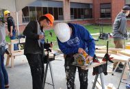 Two students working with tools, wearing helmets, cutting wood.